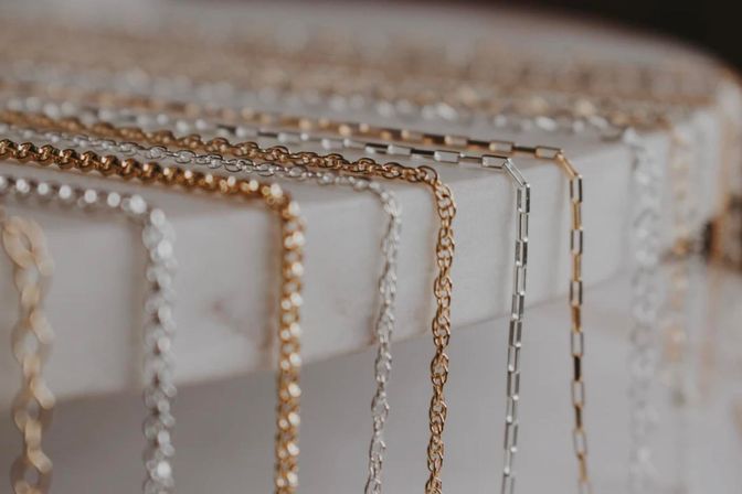 Close-up of gleaming gold and silver necklace chains draped over a white marble display edge, highlighting delicate link and box-chain styles