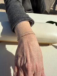 Sunlit hand resting on a cream outdoor patio cushion, gray sleeve visible and two delicate silver chain bracelets on the wrist.