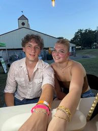 Two smiling young people at a summer county fair table with a barn-style pavilion behind them, glowing string light overhead and colorful wristbands and gold bracelets on their outstretched arms.