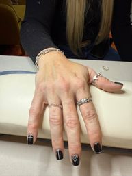 Hand with sleek black manicure featuring white minimalist lines and a V accent, wearing silver rings and a chain bracelet, resting on a salon manicure cushion.