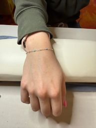 Hand resting on a manicure armrest wearing a delicate silver chain bracelet with tiny turquoise beads, pink painted nail and green sleeve.