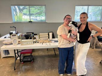 Two smiling women holding a black-and-white puppy in front of a vendor table with jewelry displays at an indoor community market with large windows.