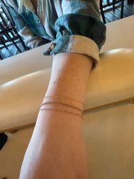 Close-up of a forearm wearing three delicate gold chain bracelets, green plaid flannel sleeve rolled up, resting on a beige cushioned armrest in an indoor setting.