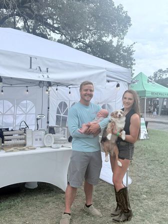 Smiling couple at an outdoor artisan market under a white canopy in a grassy park — man holding a newborn, woman holding a small brown-and-white dog, display table with handmade goods and string lights.