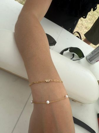 Close-up of a forearm resting on a white table wearing two delicate gold bracelets — a paperclip-style chain with a small block-letter charm and a fine chain dotted with three pearls, sunglasses and a drink can visible in a casual outdoor setting.