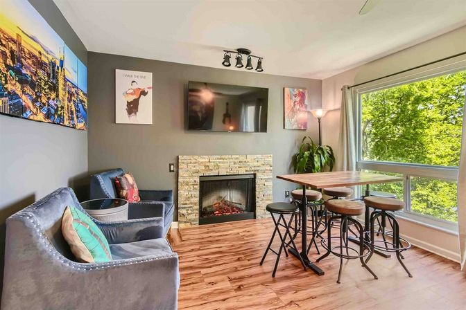 Bright modern living room and dining nook with stone fireplace, wall-mounted TV, velvet armchairs, high wooden table with stools, hardwood floors and a large window overlooking leafy trees.