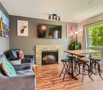 Bright modern living room and dining nook with stone fireplace, wall-mounted TV, velvet armchairs, high wooden table with stools, hardwood floors and a large window overlooking leafy trees.