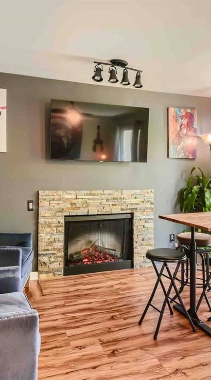 Bright modern living room and dining nook with stone fireplace, wall-mounted TV, velvet armchairs, high wooden table with stools, hardwood floors and a large window overlooking leafy trees.