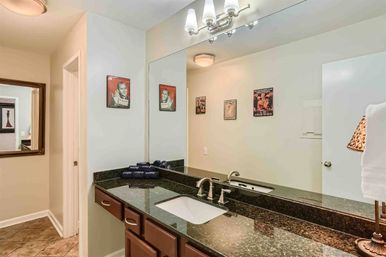Stylish residential bathroom vanity with dark granite countertop, undermount sink and brushed-nickel faucet, large wall mirror reflecting vintage poster art, folded towels, lamp and tiled floor