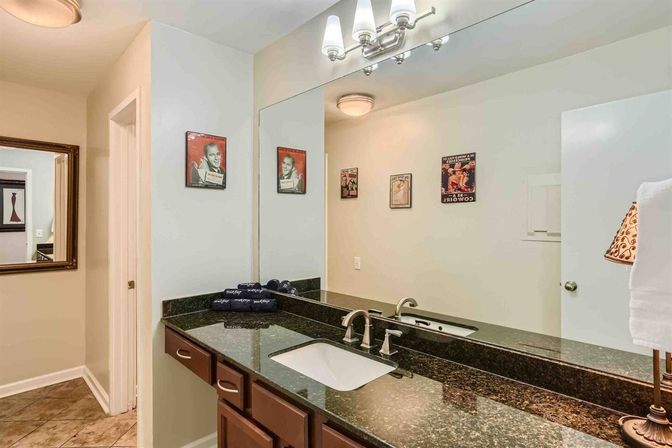 Stylish residential bathroom vanity with dark granite countertop, undermount sink and brushed-nickel faucet, large wall mirror reflecting vintage poster art, folded towels, lamp and tiled floor