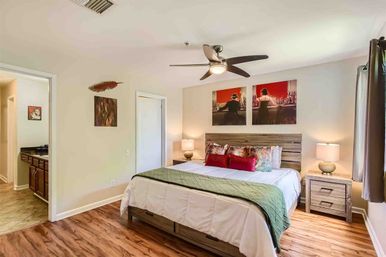 Cozy staged apartment bedroom with king bed, rustic wood headboard, green quilt and red accent pillows, matching nightstands and lamps, ceiling fan, wood-look floors, wall art and open doorway to a tiled bathroom.