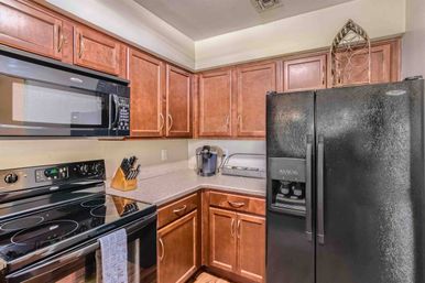 Cozy modern kitchen corner with warm wood cabinets, laminate counters, black smooth-top electric range and microwave, side-by-side fridge with water dispenser, knife block and single-serve coffee maker.