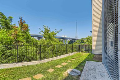 Small urban side yard with green lawn and stepping-stone path, black metal fence overlooking dense trees, a concrete highway overpass and tall radio tower in the distance, and a beige building with metal security grate at the edge