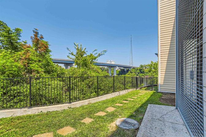 Small urban side yard with green lawn and stepping-stone path, black metal fence overlooking dense trees, a concrete highway overpass and tall radio tower in the distance, and a beige building with metal security grate at the edge