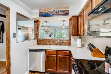 Bright open-concept kitchen with warm wood cabinets, stainless dishwasher, glass-top stove and knife block, plus a wood-framed pass-through window and pendant lights overlooking a sunlit living area and backyard view.