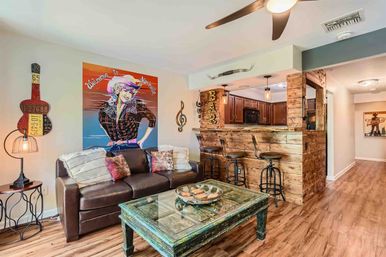 Nashville-themed living room with leather sofa, colorful "Welcome to Nashville" pop art, guitar wall decor, distressed teal coffee table, rustic wooden bar with three metal stools, pendant lights and wood floors.