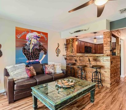Nashville-themed living room with leather sofa, colorful "Welcome to Nashville" pop art, guitar wall decor, distressed teal coffee table, rustic wooden bar with three metal stools, pendant lights and wood floors.