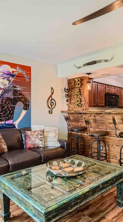 Nashville-themed living room with leather sofa, colorful "Welcome to Nashville" pop art, guitar wall decor, distressed teal coffee table, rustic wooden bar with three metal stools, pendant lights and wood floors.
