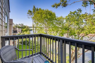 Cozy riverfront balcony with wicker chair and gray wooden railing overlooking leafy trees, calm river and distant bridge on a sunny day.