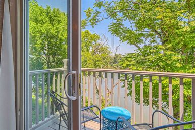 Sunny balcony seen through a sliding glass door with metal patio chairs and a blue side table, overlooking lush green trees and a calm river view.