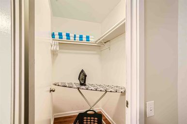 Small home linen closet with an ironing board and black iron on a patterned cover, blue-and-white striped folded towels on a shelf, white hangers and a laundry basket on hardwood floor.