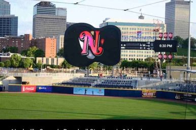 Downtown baseball park outfield with green grass, a giant black scoreboard showing a pink 'N' logo and 6:30 PM, bleachers and city skyline in the background.