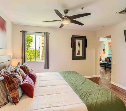 Cozy bright apartment bedroom with king bed, layered colorful pillows and green quilted runner, modern ceiling fan, hardwood floors, wall art and TV, plus a window view of trees and an open doorway to the living area.