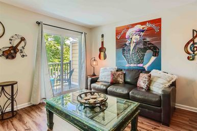 Cozy living room with dark leather sofa and textured pillows, colorful ‘Welcome to Nashville’ wall art, glass-top rustic coffee table, musical wall decor, sliding glass door to a sunlit balcony and hardwood floors.