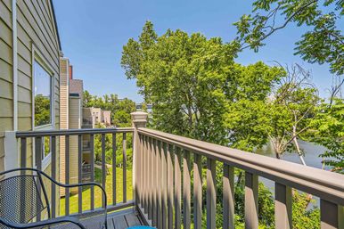 Sunny riverside balcony with a metal chair and painted railing overlooking lush green trees and a calm river.