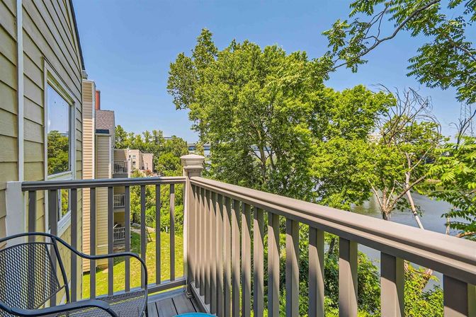 Sunny riverside balcony with a metal chair and painted railing overlooking lush green trees and a calm river.