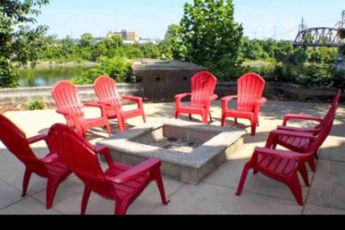 Eight red Adirondack chairs arranged around a square concrete fire pit on a sunny riverside patio with greenery and a metal bridge visible in the background.