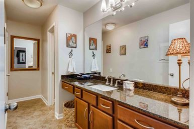 Music-themed bathroom vanity with dark granite countertop, undermount sink, warm wood cabinets, large mirror, patterned lamp, treble-clef towel hook, wicker basket and tile floor.