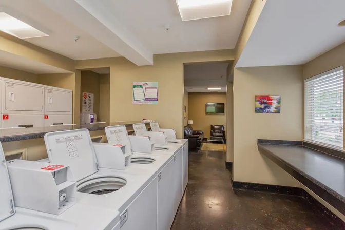 Shared apartment laundry room with a row of open top-loading washers, stacked dryers, long folding counter by a window with blinds, and a small lounge area with chairs and TV — bright beige interior ready for laundry day.