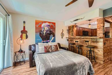 Cozy Nashville-themed studio apartment with a pull-out bed on a leather sofa, colorful pop-art "Welcome to Nashville" poster above, rustic reclaimed-wood bar with three industrial stools, sliding glass door, and warm pendant lighting.