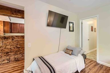 Small cozy bedroom with single bed dressed in white linens and striped throw, wall-mounted flat-screen TV, rustic reclaimed wood accent paneling, hardwood floors and open doorway to a hallway