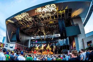 Crowded outdoor amphitheater with a large curved roof, band performing on a brightly lit stage with rigging and colorful spotlights, lively audience enjoying a live music concert.