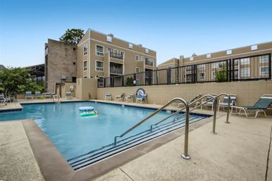 Rectangular outdoor swimming pool with floaties, lounge chairs and stainless handrails in a beige multi-story apartment complex courtyard under a clear blue sky.
