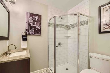 Compact residential bathroom with corner glass shower, white subway tile and horizontal mosaic accent, chrome fixtures, stone-top vanity, toilet, and black-and-white wall art on neutral beige walls.