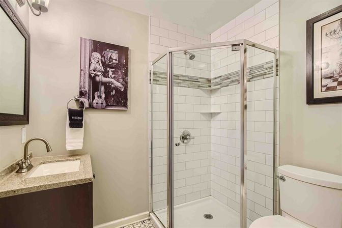 Compact residential bathroom with corner glass shower, white subway tile and horizontal mosaic accent, chrome fixtures, stone-top vanity, toilet, and black-and-white wall art on neutral beige walls.