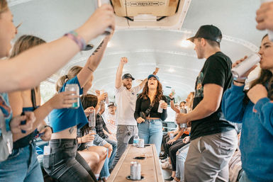 Young adults cheering and holding drinks inside a lively party bus interior during a group road-trip outing