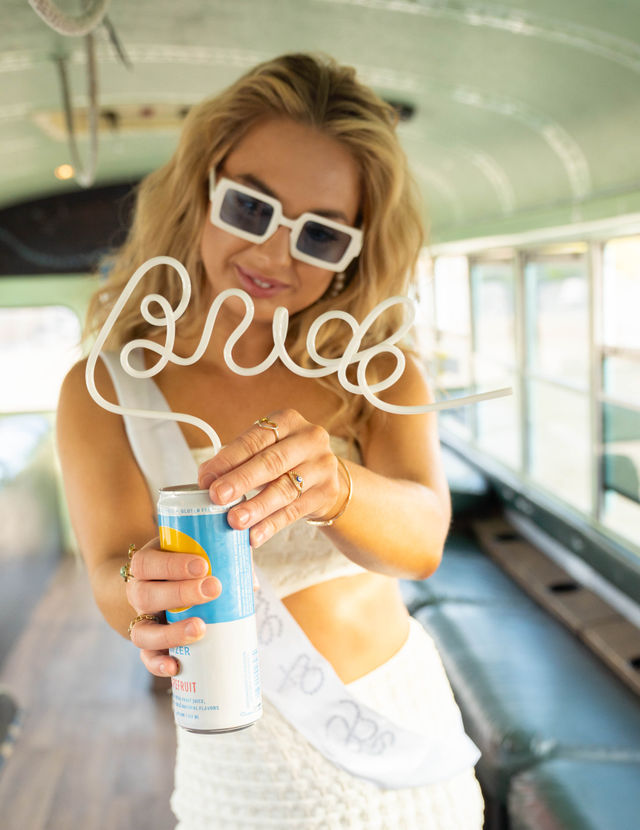 Bride-to-be in a white sash and sunglasses inside a vintage green party bus, smiling as she opens a canned drink with a playful curly straw that spells "bride".