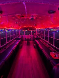 Vibrant pink-lit party bus interior with mirrored disco ball, multicolored string lights, long black bench seating along both sides, wooden aisle floor and a small center table — festive nighttime vibe.