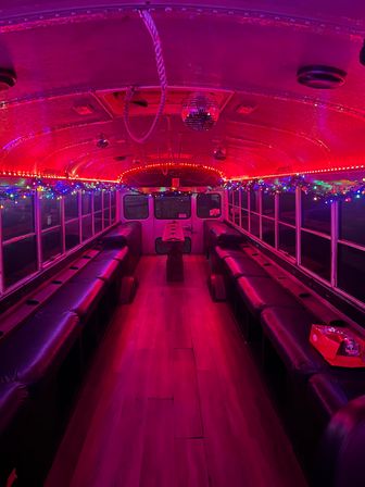 Vibrant pink-lit party bus interior with mirrored disco ball, multicolored string lights, long black bench seating along both sides, wooden aisle floor and a small center table — festive nighttime vibe.