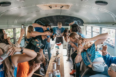Group of friends partying inside a converted school bus, chugging canned drinks and playing around a long center table strewn with red cups and spilled liquid.