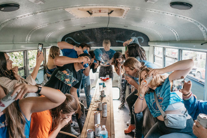 Group of friends partying inside a converted school bus, chugging canned drinks and playing around a long center table strewn with red cups and spilled liquid.