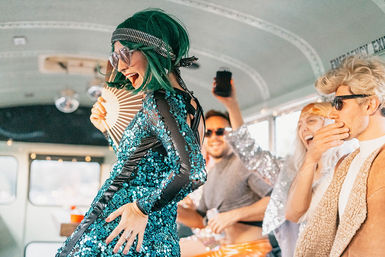 Group of friends partying inside a vintage bus — woman in green wig and teal sequin dress dancing as others cheer, laugh, and take photos.