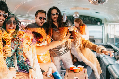Group of people in glittery retro outfits and faux fur coats partying on a decorated party bus, laughing and raising drink cans under a disco ball.