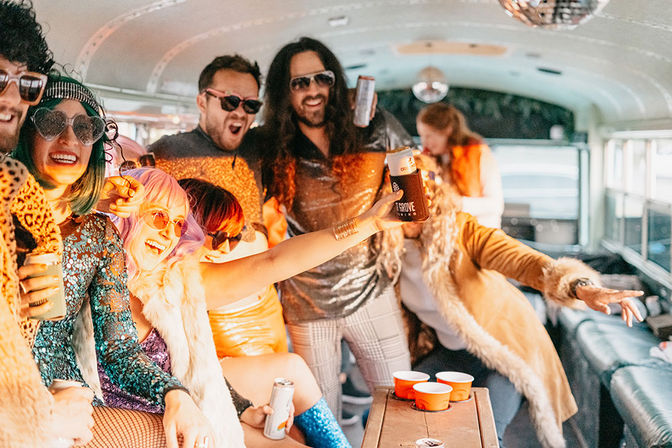 Group of people in glittery retro outfits and faux fur coats partying on a decorated party bus, laughing and raising drink cans under a disco ball.