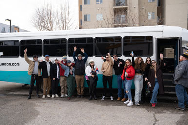 Group of friends in winter jackets posing and cheering beside a white-and-teal shuttle bus parked in an urban apartment lot, many holding drinks and raising their hands.
