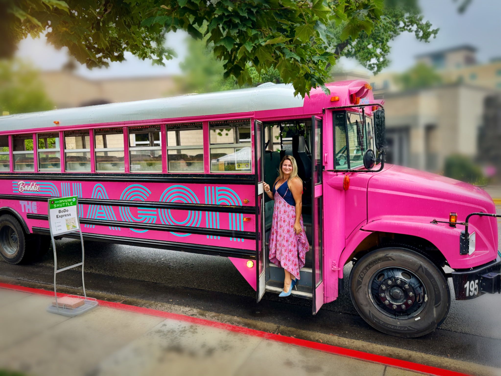 Bright pink vintage-style shuttle bus parked on a downtown curb under leafy trees, a smiling woman in a floral dress stepping out onto wet pavement.